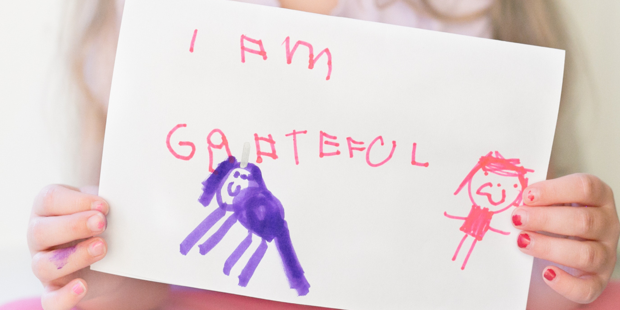 Tiny hands with red chipped nail polish hold a rectangle of white paper. Written in pink marker in a child's handwriting is 