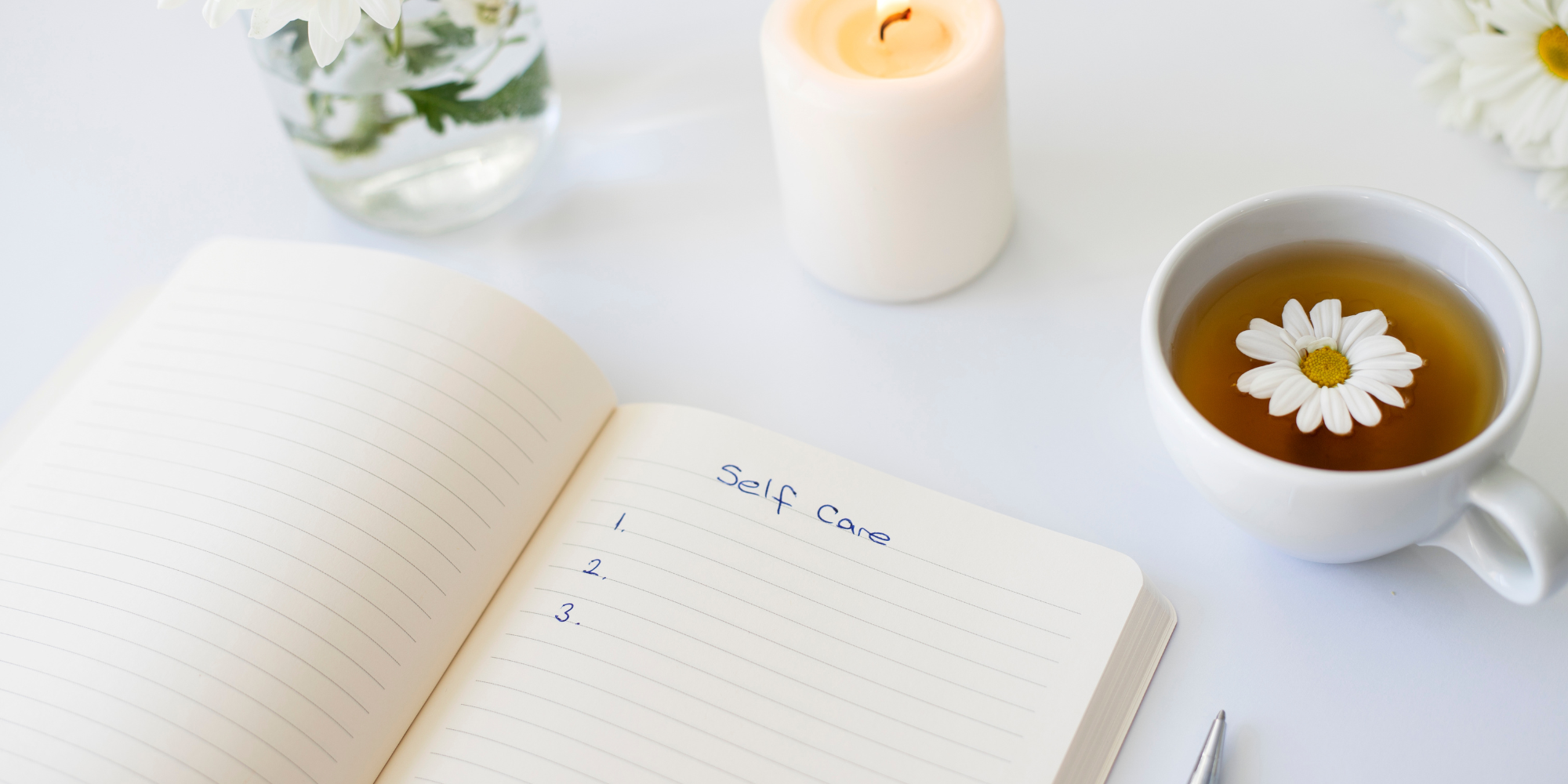 Journal open with the text "self care" at the top with numbered blanks below. There is a candle, a vase with flowers and cup of tea on the white table as well. 