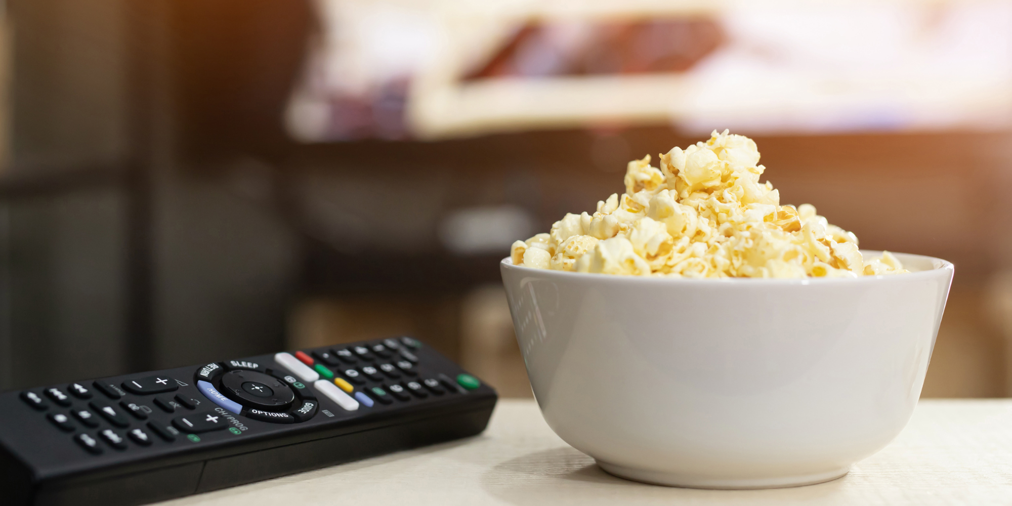 a TV remote next to a bowl of popcorn. Blurred in the distance, you can see a turned-on TV screen.