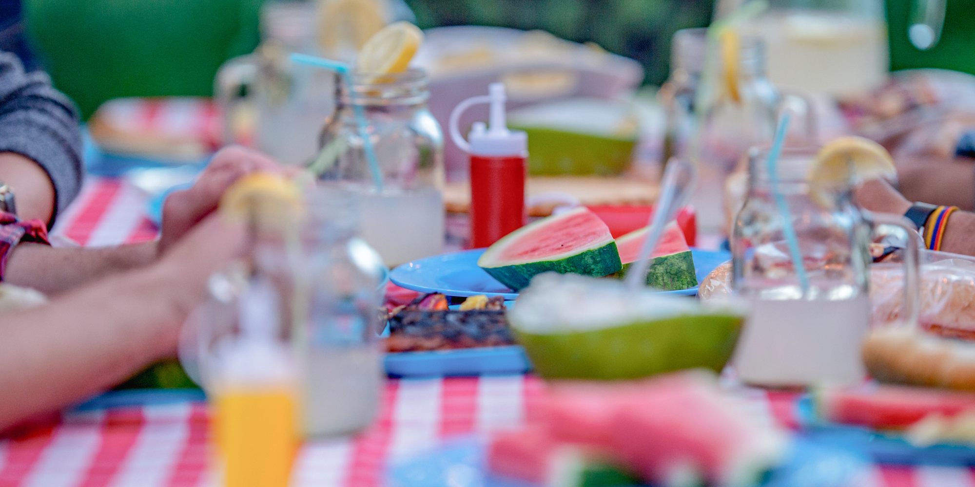 Table at a family cookout/barbeque. The table has a red and white checkered tablecloth, blue plates, watermelon, some grilled vegetables, mason jars filled with lemonade and a catchup bottle in the distance.  