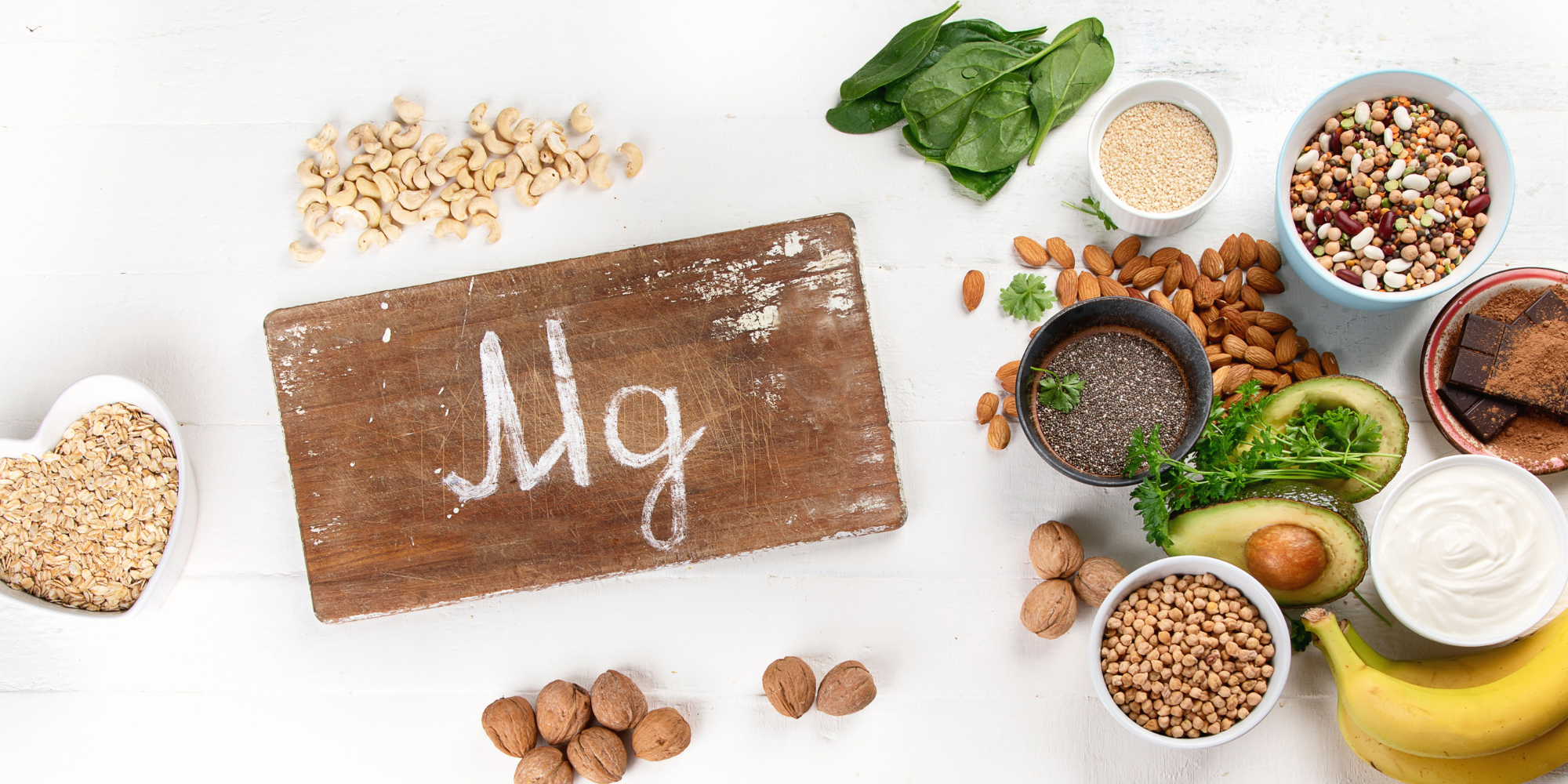 Various magnesium rich foods including dark chocolate, nuts, beans, spinach, chia seeds, and avocado, all around a sign on a cutting board that reads "Mg". 