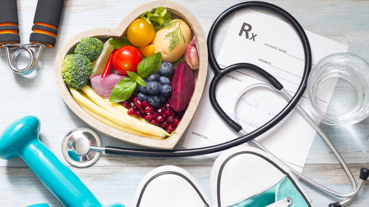 At the top left, there is a heart shaped bowl with fresh whole foods in it. At the bottom of the image, there are exercise shoes and weights. Intertwined between the bowl and exercise equipment there is a black stethoscope .